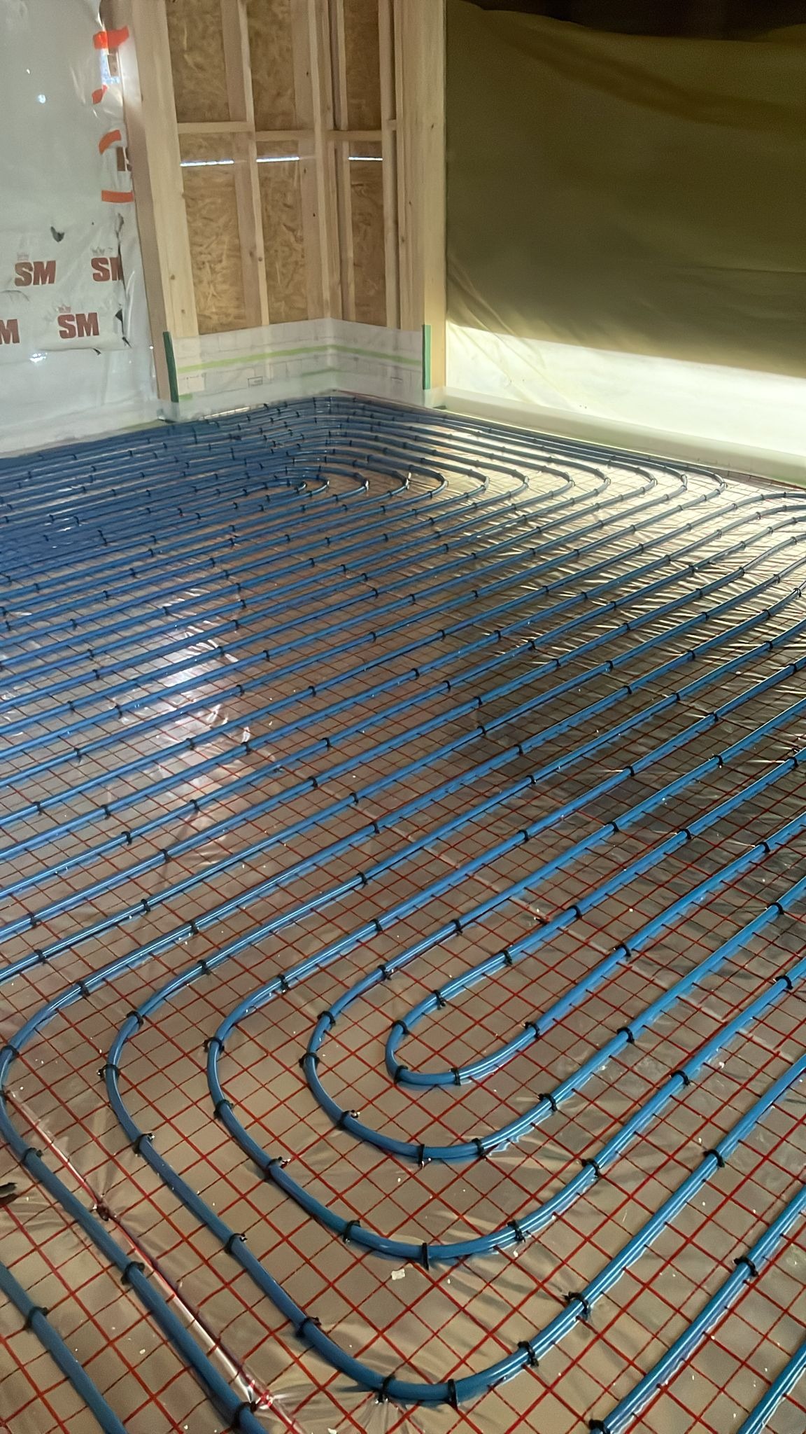 Niebieskie rury ogrzewania podłogowego ułożone na czerwonej siatce w surowym wnętrzu, widoczne elementy konstrukcyjne ścian i folia ochronna.