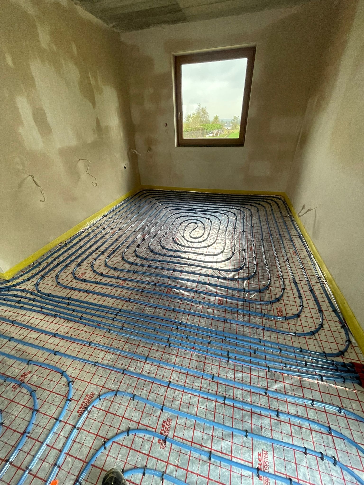 Niebieskie rury ogrzewania podłogowego ułożone spiralnie na siatce w surowym pomieszczeniu z oknem, zabezpieczone taśmą przy ścianach.