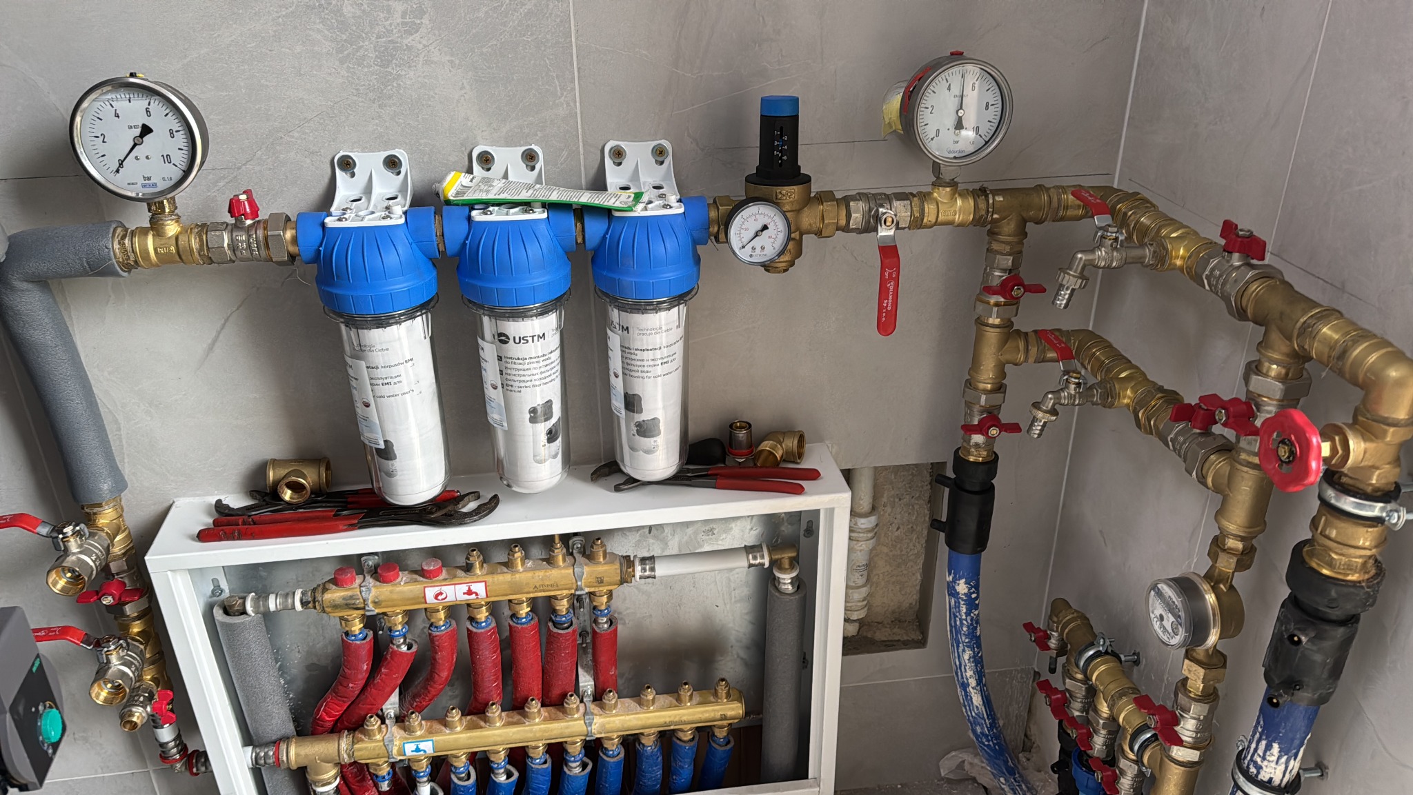 Rozbudowana instalacja hydrauliczna z filtrami wody, manometrami i rozdzielaczem ogrzewania podłogowego w szafce, widoczne narzędzia monterskie.
