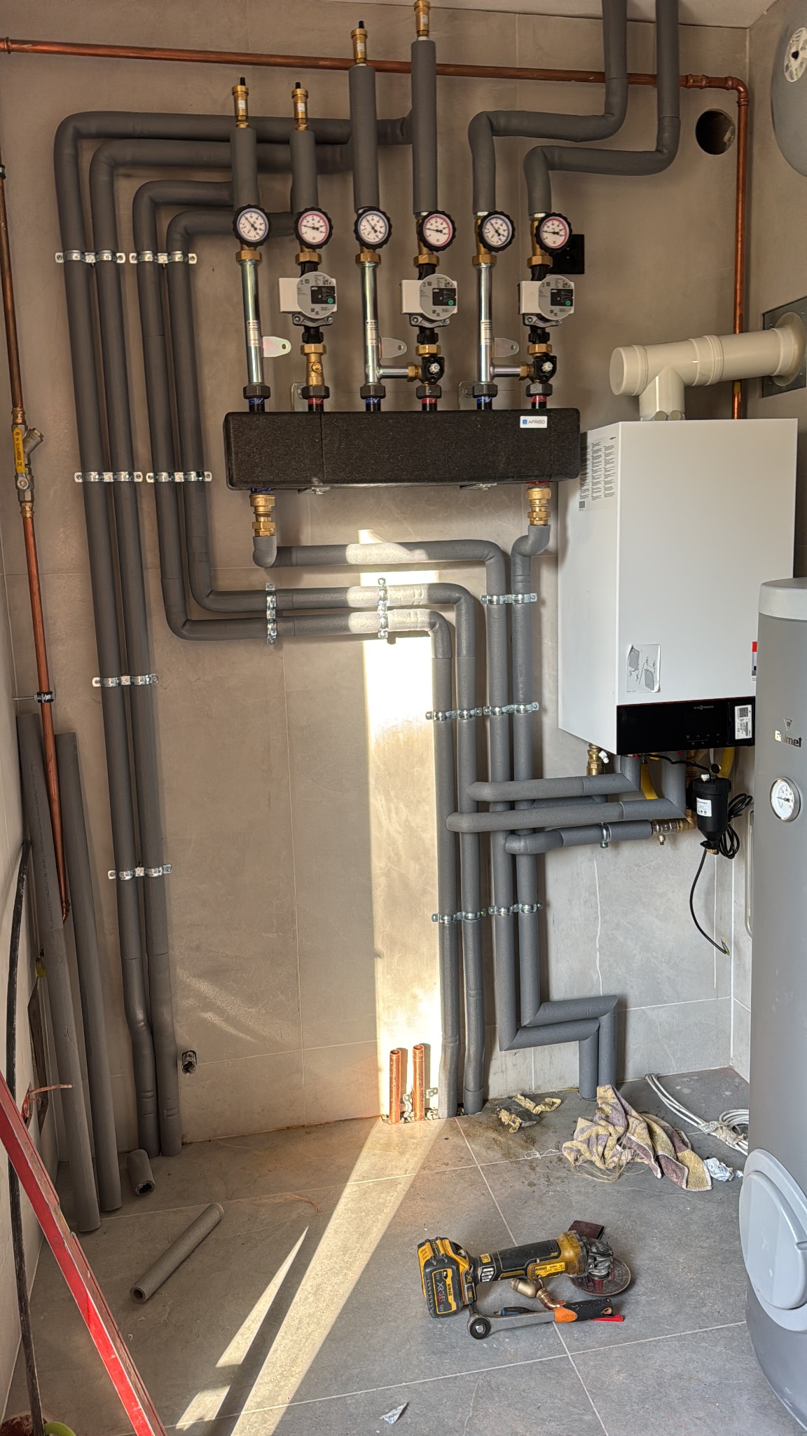 Rozbudowana instalacja grzewcza z rozdzielaczem, rurami w izolacji, kotłem gazowym i zasobnikiem ciepłej wody. Na podłodze narzędzia: szlifierka kątowa akumulatorowa DeWalt i klucz.