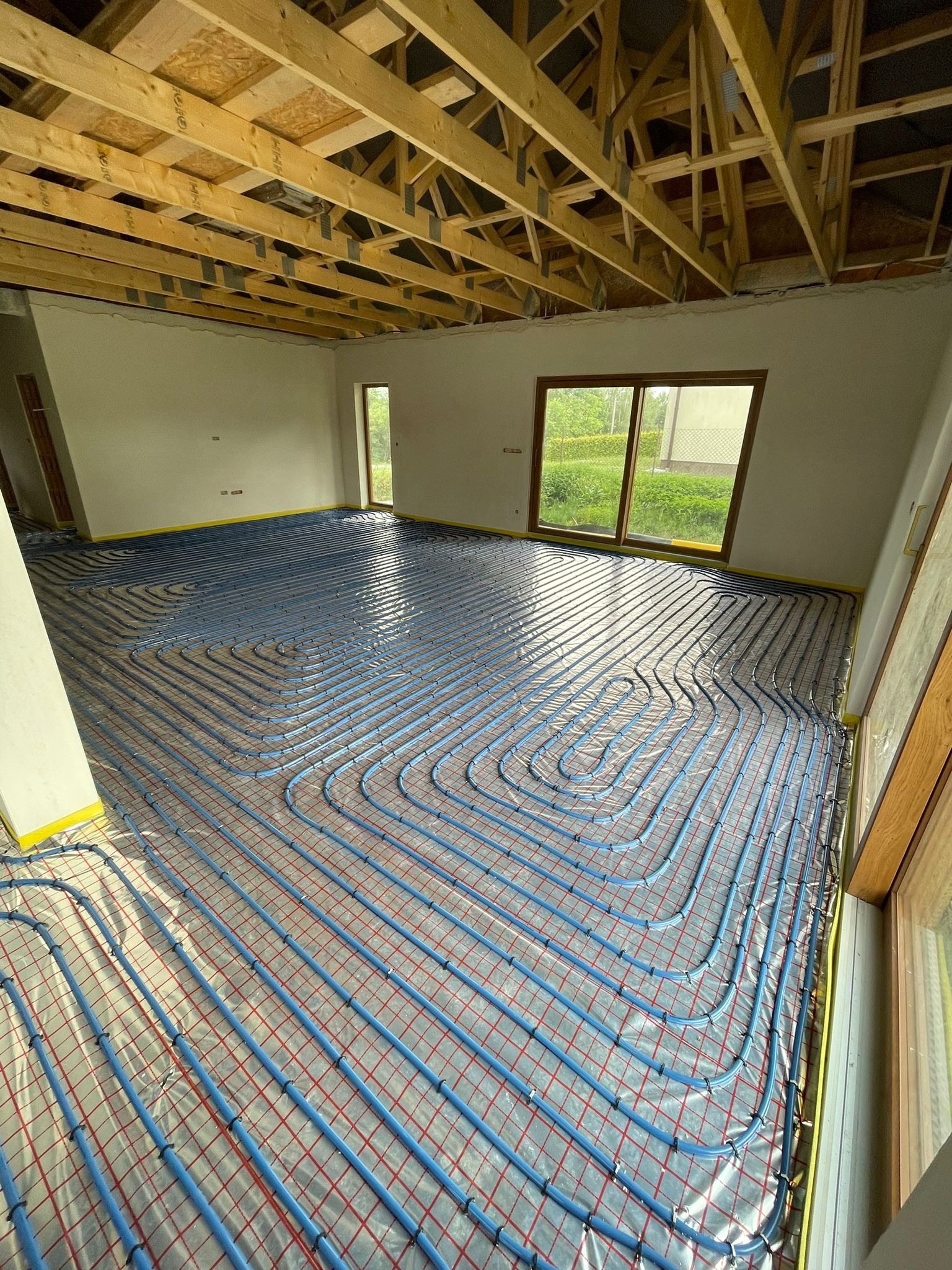 Niebieskie rury ogrzewania podłogowego ułożone na folii z czerwoną siatką w surowym wnętrzu z drewnianą konstrukcją stropu i oknami z widokiem na zieleń.