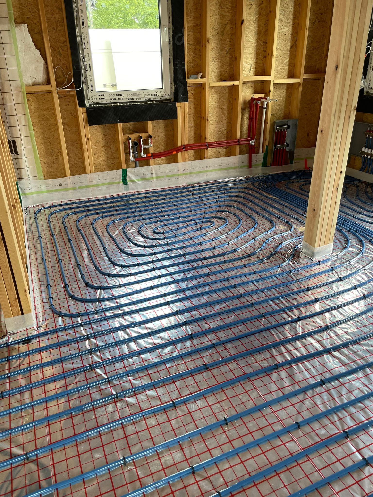 Instalacja ogrzewania podłogowego w nowym budynku, widoczne niebieskie rury układane spiralnie na srebrnej folii z czerwoną siatką, rozdzielacz na ścianie, surowa konstrukcja drewniana budynku.