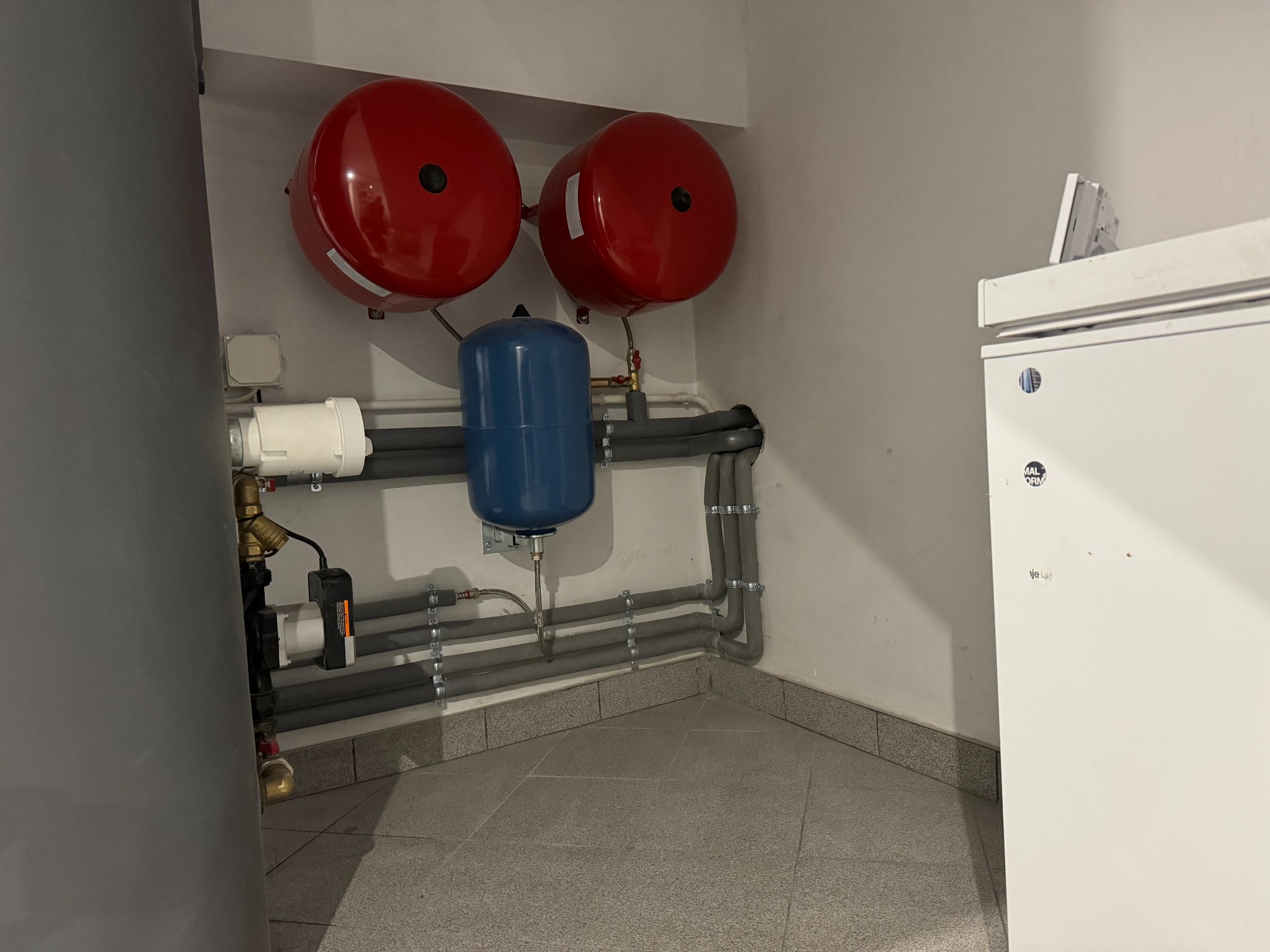 Dwa czerwone zbiorniki ciśnieniowe, niebieski zbiornik wyrównawczy i szare rury instalacji w pomieszczeniu technicznym z szarą podłogą z płytek.