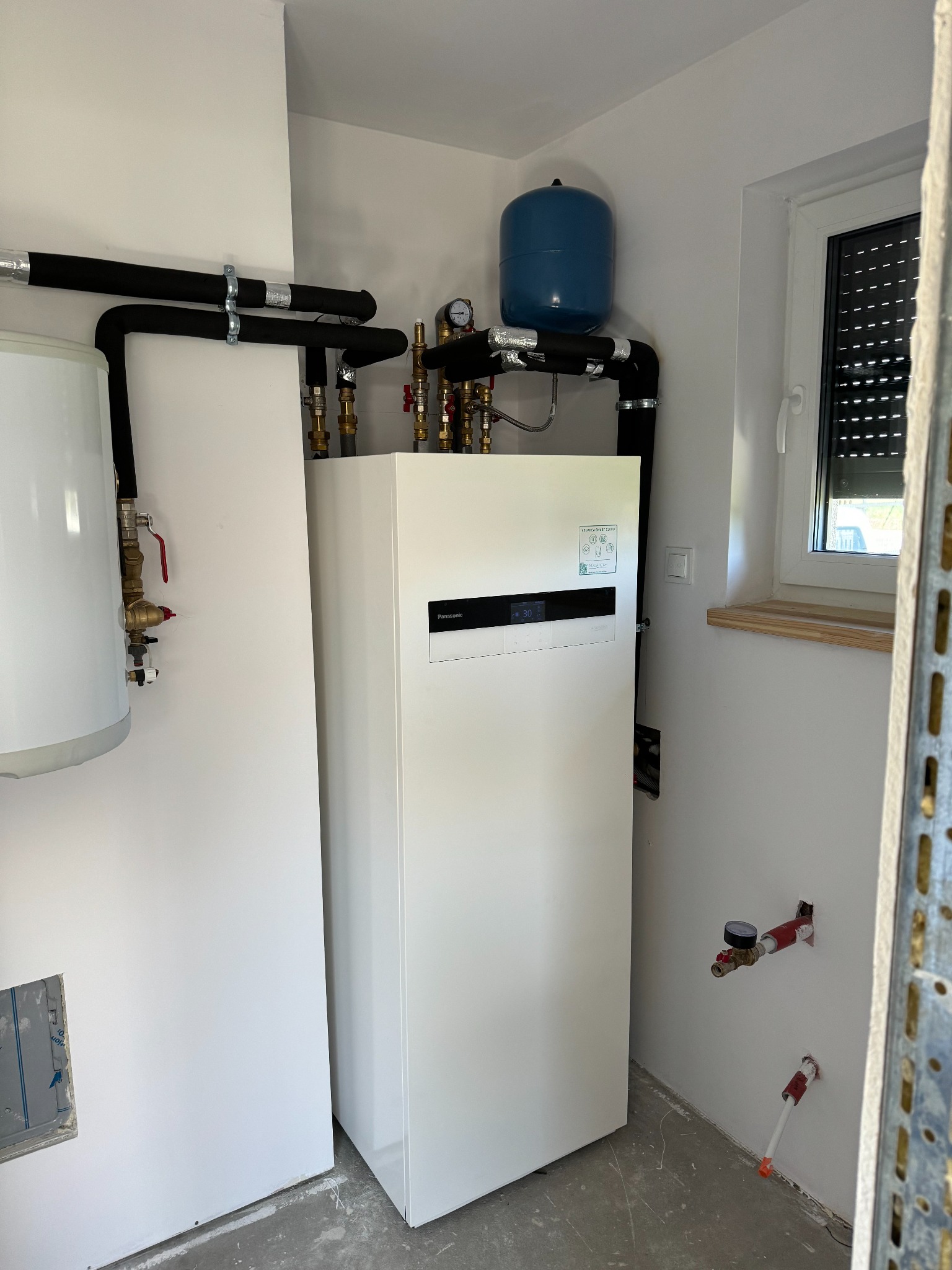 Nowoczesna, biała pompa ciepła Panasonic Aquarea Smart Cloud, z podłączonym zbiornikiem wyrównawczym w kolorze niebieskim i orurowaniem izolowanym czarną pianką, zamontowana w pomieszczeniu z oknem...