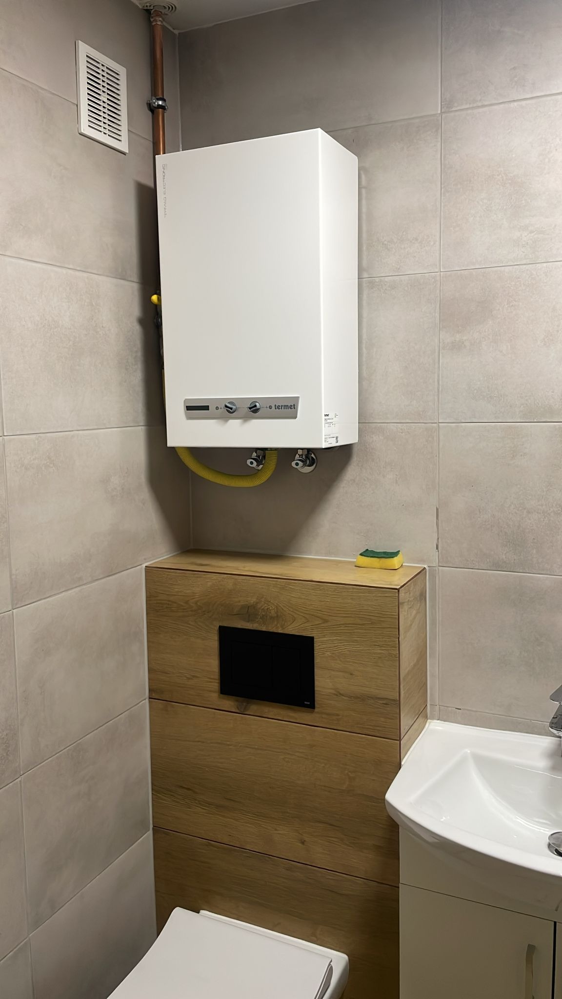 Biały piec gazowy Termet zamontowany na szarej, kafelkowanej ścianie nad zabudową WC z drewnopodobną okleiną i żółtą rurą gazową, widoczny fragment umywalki.
