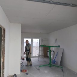 Prof Bud - Montaż sufitu podwieszanego z płyty karton gips, widoczny pracownik w kombinezonie, podnośnik do płyt, drabina i narzędzia w surowym wnętrzu mieszkania.