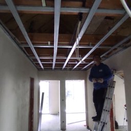 Prof Bud - Montaż metalowej konstrukcji sufitu podwieszanego w trakcie prac wykończeniowych, widoczny pracownik na drabinie w pomieszczeniu z surowymi ścianami i instalacjami.