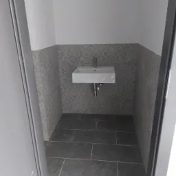 Mała łazienka z kwadratową umywalką, szarymi płytkami z wzorem na ścianie i ciemnymi płytkami na podłodze, widok zza otwartych drzwi.