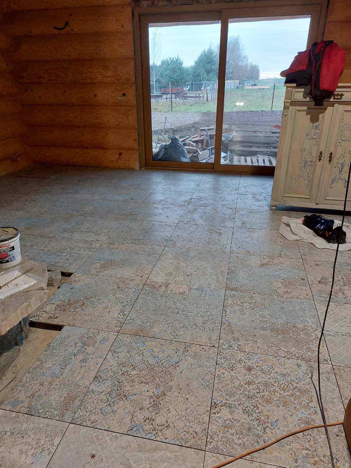 Układanie płytek podłogowych w drewnianym domu, widoczne niedokończone fragmenty, klej i narzędzia, okno z widokiem na posesję.