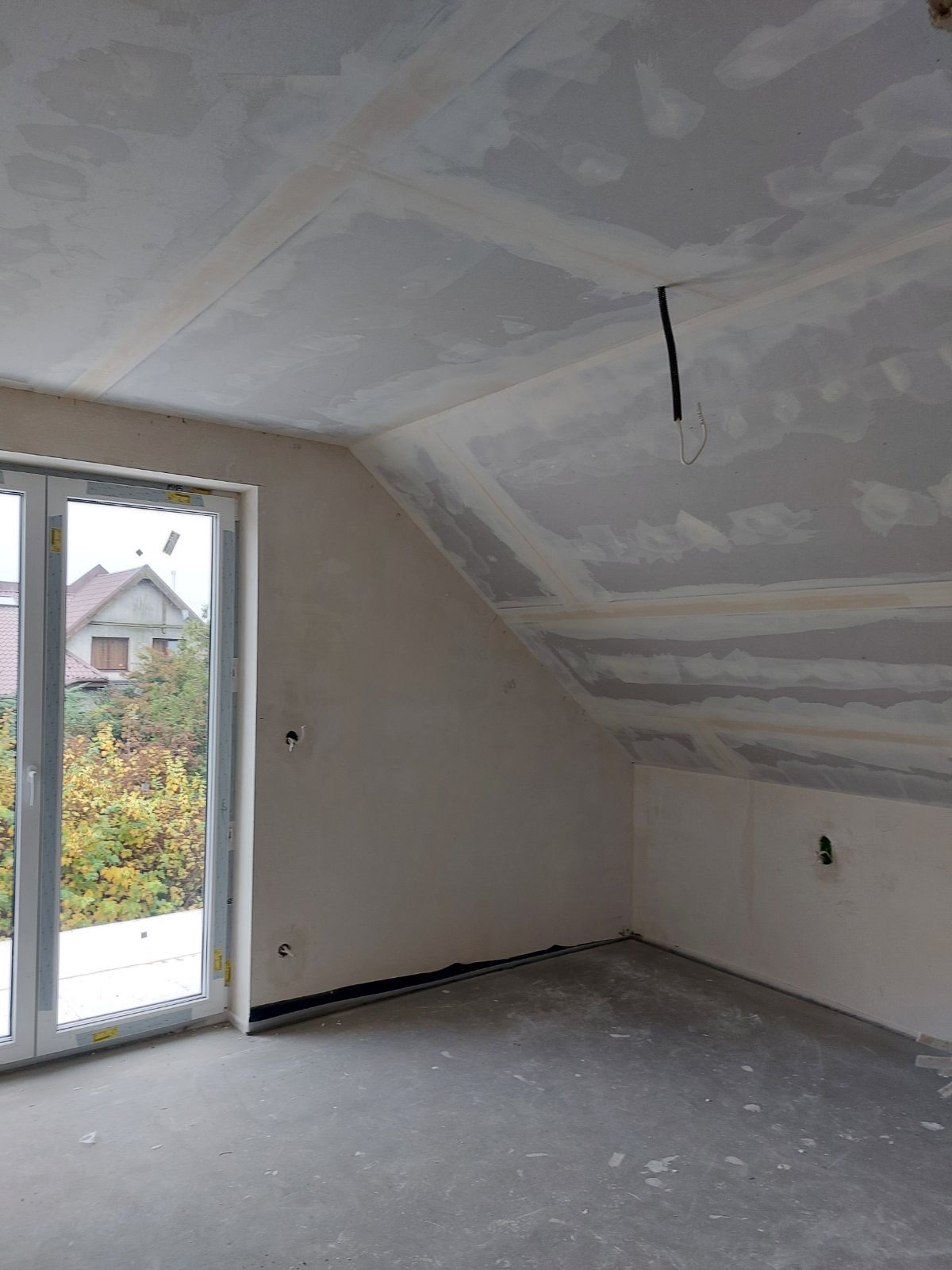 Poddasze w trakcie prac wykończeniowych, widoczne płyty kartonowo-gipsowe na skosach i suficie, zaszpachlowane łączenia, okno dachowe z widokiem na jesienne drzewa.