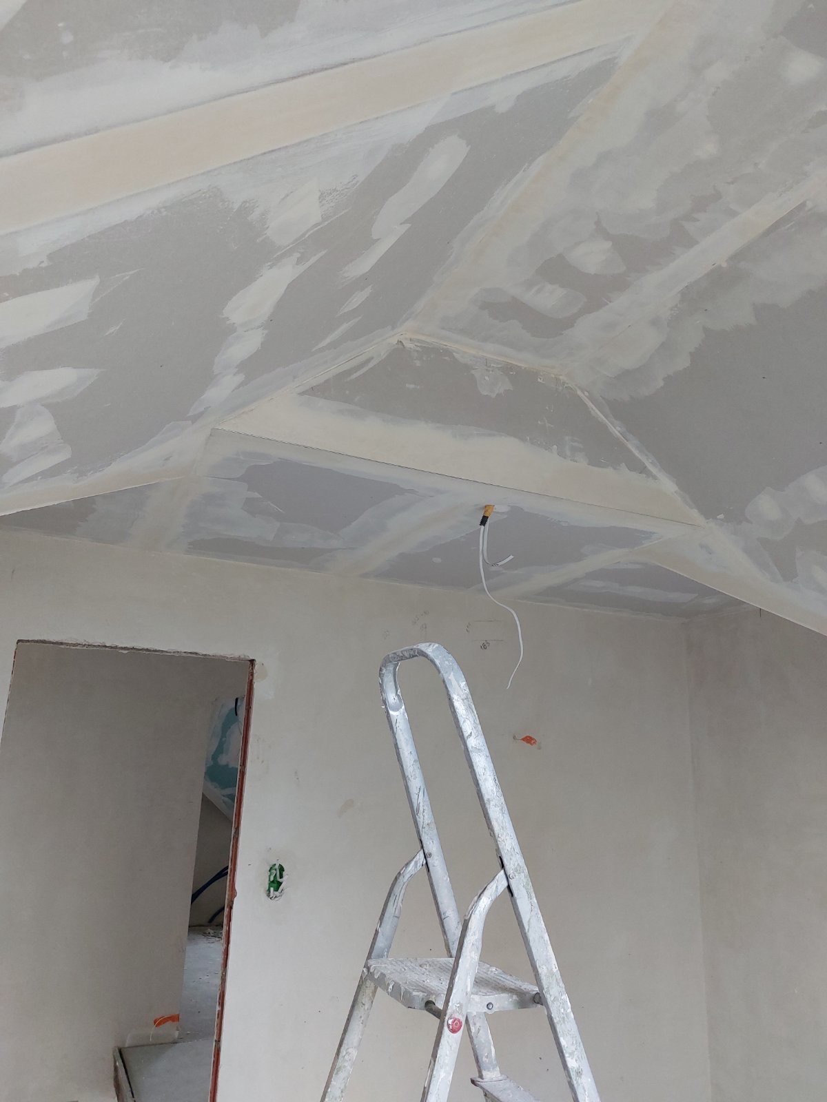 Skośny sufit z płyt kartonowo-gipsowych w trakcie szpachlowania, widoczna aluminiowa drabina i wystające kable, fragment otworu drzwiowego w tle.