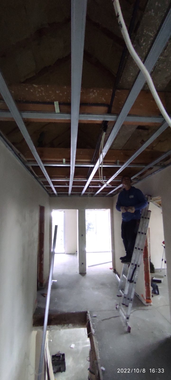 Montaż metalowej konstrukcji sufitu podwieszanego w trakcie prac wykończeniowych, widoczny pracownik na drabinie w pomieszczeniu z surowymi ścianami i instalacjami.