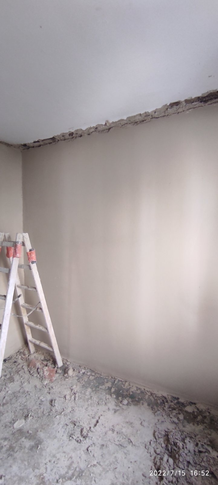 Przygotowanie do malowania: Ściana po usunięciu starej farby, widoczne ślady po skrobaniu, drabina oparta o ścianę, gruz na podłodze, narożnik sufitu z widocznymi uszkodzeniami.