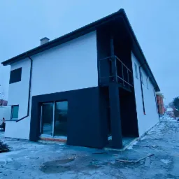 Dwukondygnacyjny dom z nową, dwukolorową elewacją (biały i grafitowy), balkonem i dużymi oknami, widoczny w zimowej scenerii z resztkami śniegu na ziemi.