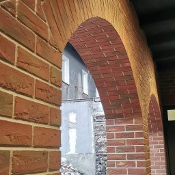Kowbud - Junior - Nowa elewacja z cegły klinkierowej tworząca łukowate przejście, widok na gruzowisko i fragment budynku w tle.