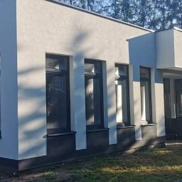 Kowbud - Junior - Nowoczesny budynek z szarą, teksturowaną elewacją i czarnymi obramowaniami okien, otoczony zielenią i drzewami.