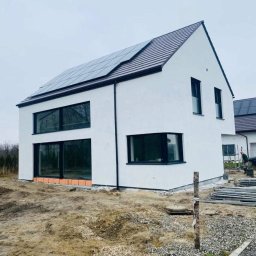 Kowbud - Junior - Nowoczesny, dwukondygnacyjny dom z białymi ścianami i ciemną stolarką okienną, panele słoneczne na dachu, teren wokół domu w trakcie zagospodarowywania.