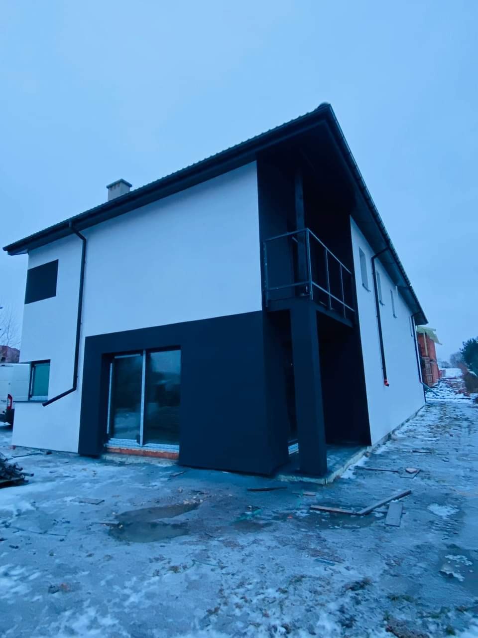 Dwukondygnacyjny dom z nową, dwukolorową elewacją (biały i grafitowy), balkonem i dużymi oknami, widoczny w zimowej scenerii z resztkami śniegu na ziemi.