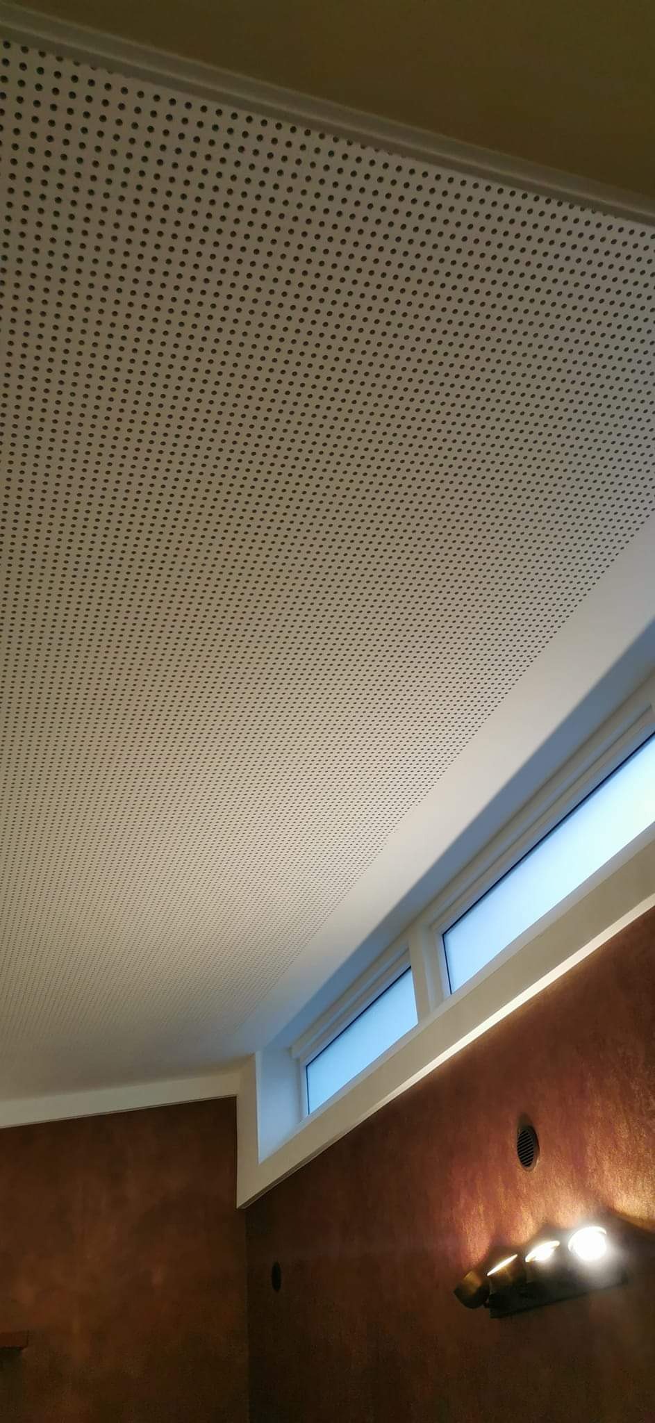 Sufit podwieszany z perforacją, widok z dołu, brązowa ściana z oknem w tle, lampa ścienna.