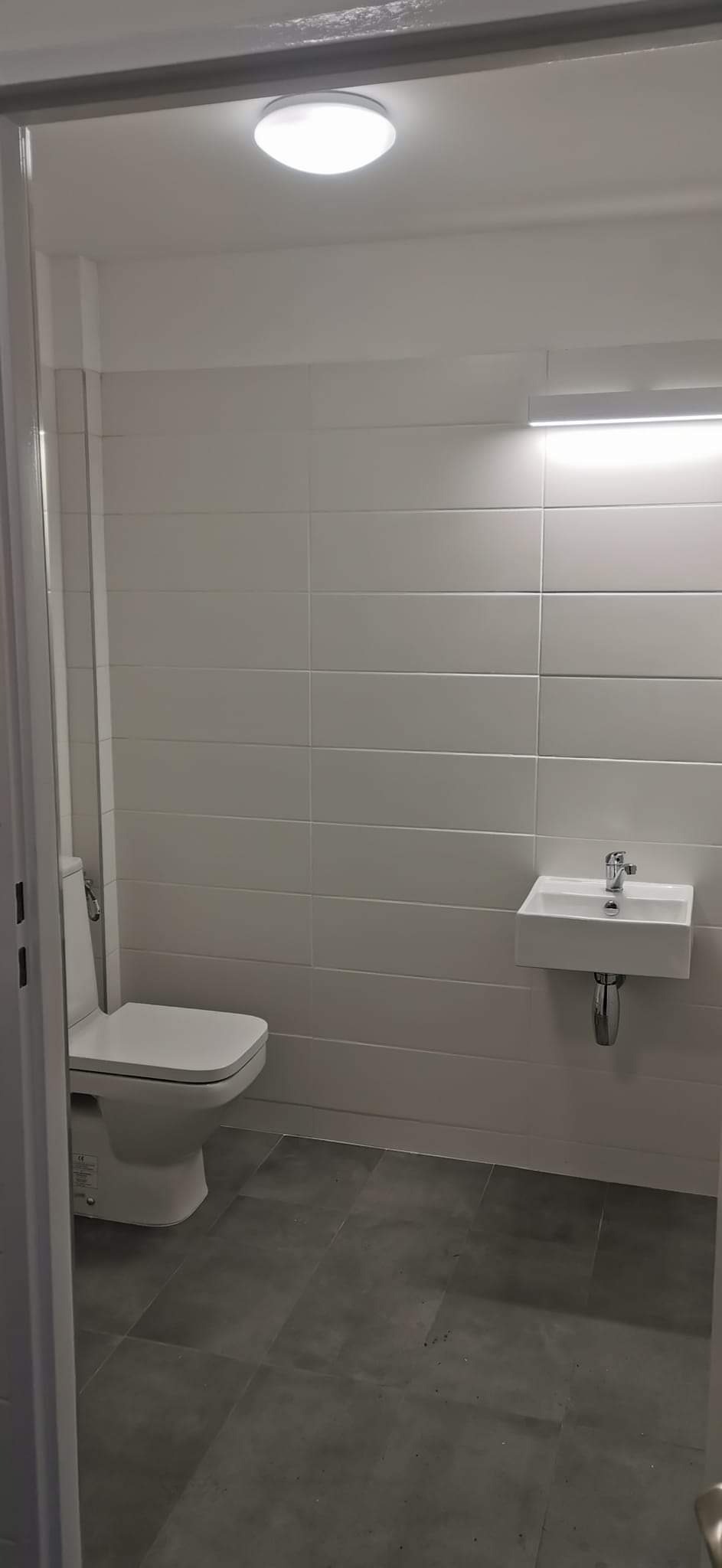 Wykończona łazienka w minimalistycznym stylu: białe płytki ścienne, podwieszana umywalka z chromowaną baterią i toaleta, szara podłoga z płytek imitujących kamień, oświetlenie sufitowe i listwa LED.