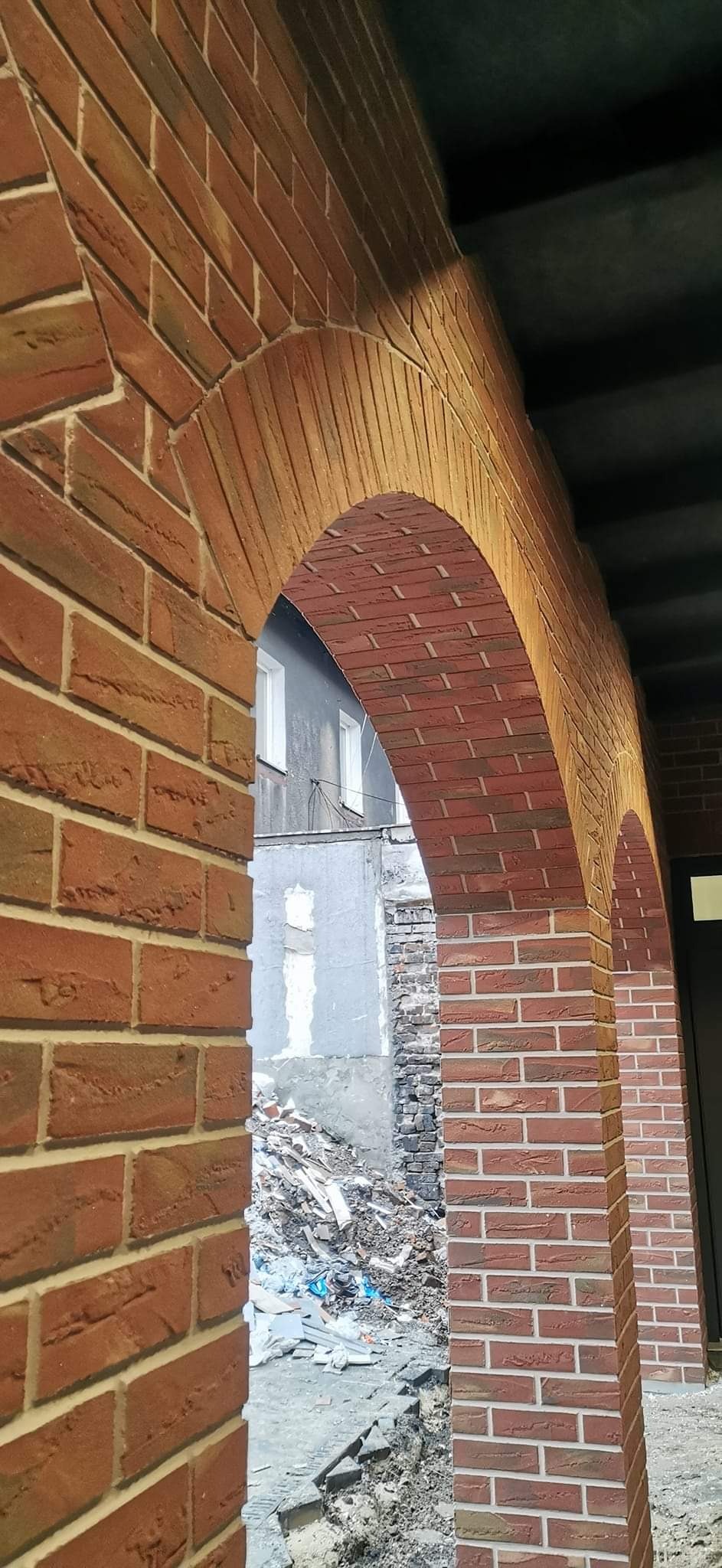 Nowa elewacja z cegły klinkierowej tworząca łukowate przejście, widok na gruzowisko i fragment budynku w tle.