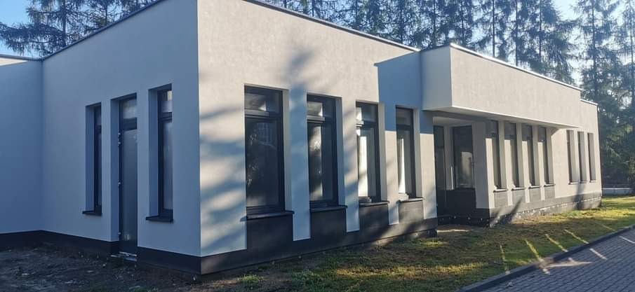 Nowoczesny budynek z szarą, teksturowaną elewacją i czarnymi obramowaniami okien, otoczony zielenią i drzewami.