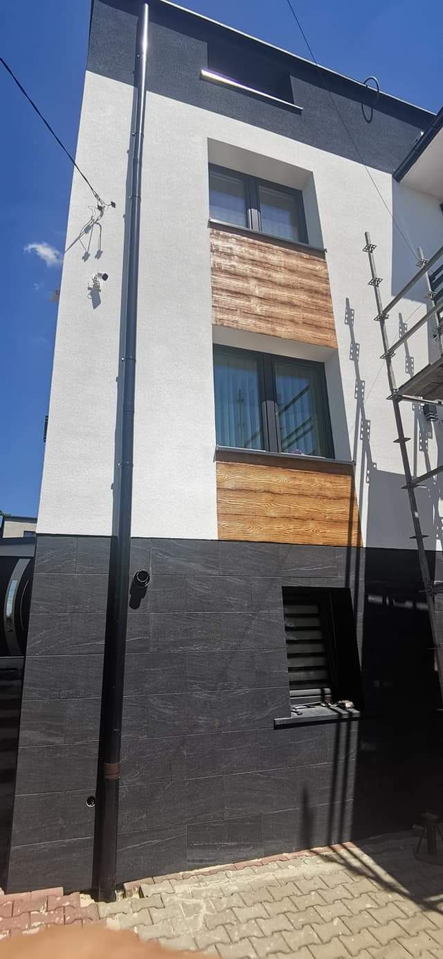 Dwukondygnacyjny budynek z elewacją w kolorach białym, szarym i imitującym drewno, z widoczną rynną i fragmentem rusztowania.