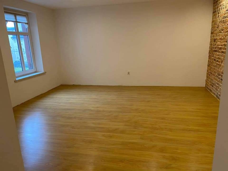 Pusty pokój po remoncie z panelami podłogowymi w odcieniach brązu, białe ściany i fragment ściany z cegły, okno z widokiem na zieleń za oknem.