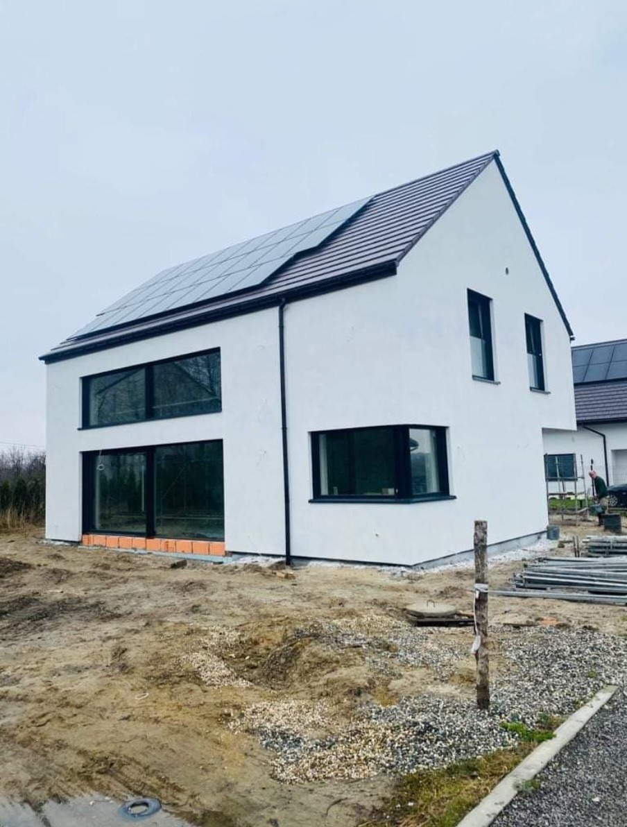 Nowoczesny, dwukondygnacyjny dom z białymi ścianami i ciemną stolarką okienną, panele słoneczne na dachu, teren wokół domu w trakcie zagospodarowywania.