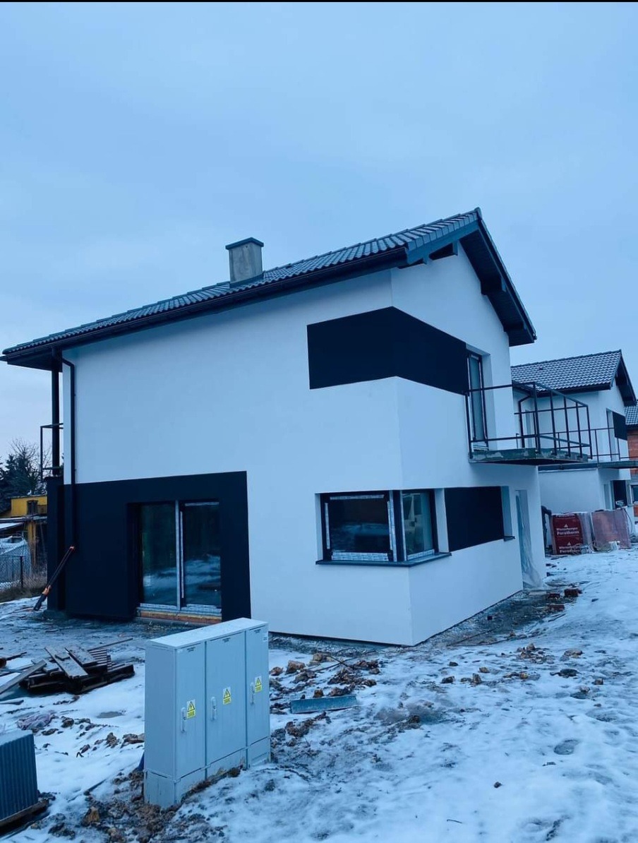 Nowoczesny dom jednorodzinny z białą elewacją i czarnymi akcentami w zimowej scenerii, widoczna skrzynka elektryczna i porozrzucane cegły.