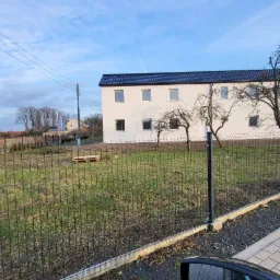 Dwukondygnacyjny, biały dom z ciemnym dachem widziany zza metalowego ogrodzenia, z fragmentem lusterka samochodowego w prawym dolnym rogu kadru.