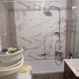 Ukończona łazienka z marmurowymi płytkami, widoczna kabina prysznicowa, umywalka z szafką, pralka z miskami, suszarka podsufitowa i oświetlenie.