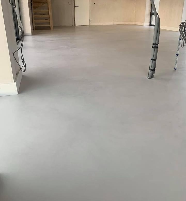 Jasnoszara, gładka podłoga w minimalistycznym wnętrzu, widoczne pionowe rury instalacyjne przy ścianie, schody w tle.