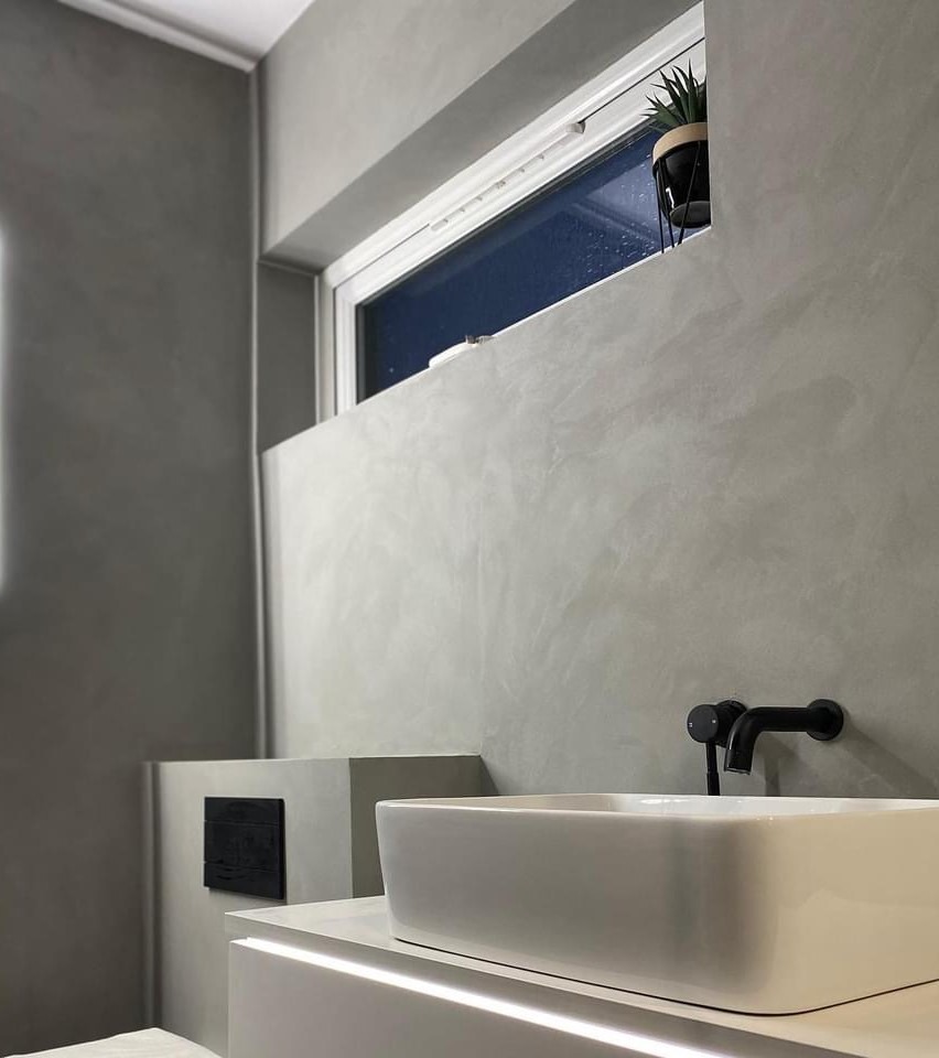 Minimalistyczna łazienka z szarą posadzką żywiczną na ścianach, białą umywalką nablatową, czarną armaturą i rośliną w doniczce w oknie.