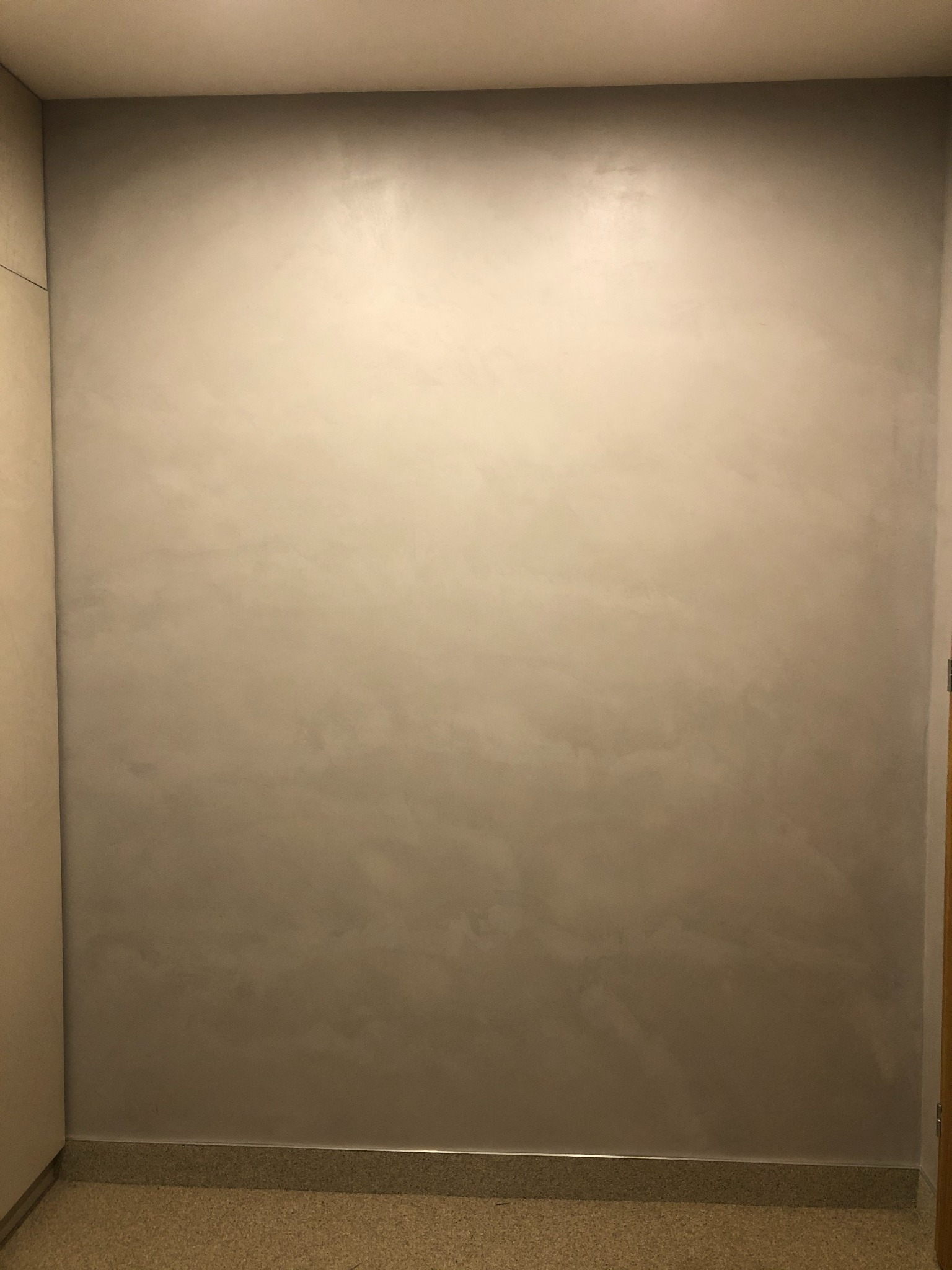 Gładka, beżowa ściana z efektem cieniowania, wykończona przy podłodze listwą przypodłogową, oświetlona od góry.