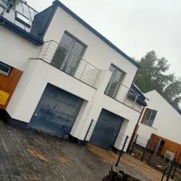 Nowoczesne domy szeregowe z balkonami i garażami, wykończone w bieli z elementami drewna, widok z zewnątrz na kostkę brukową i fragment placu budowy.