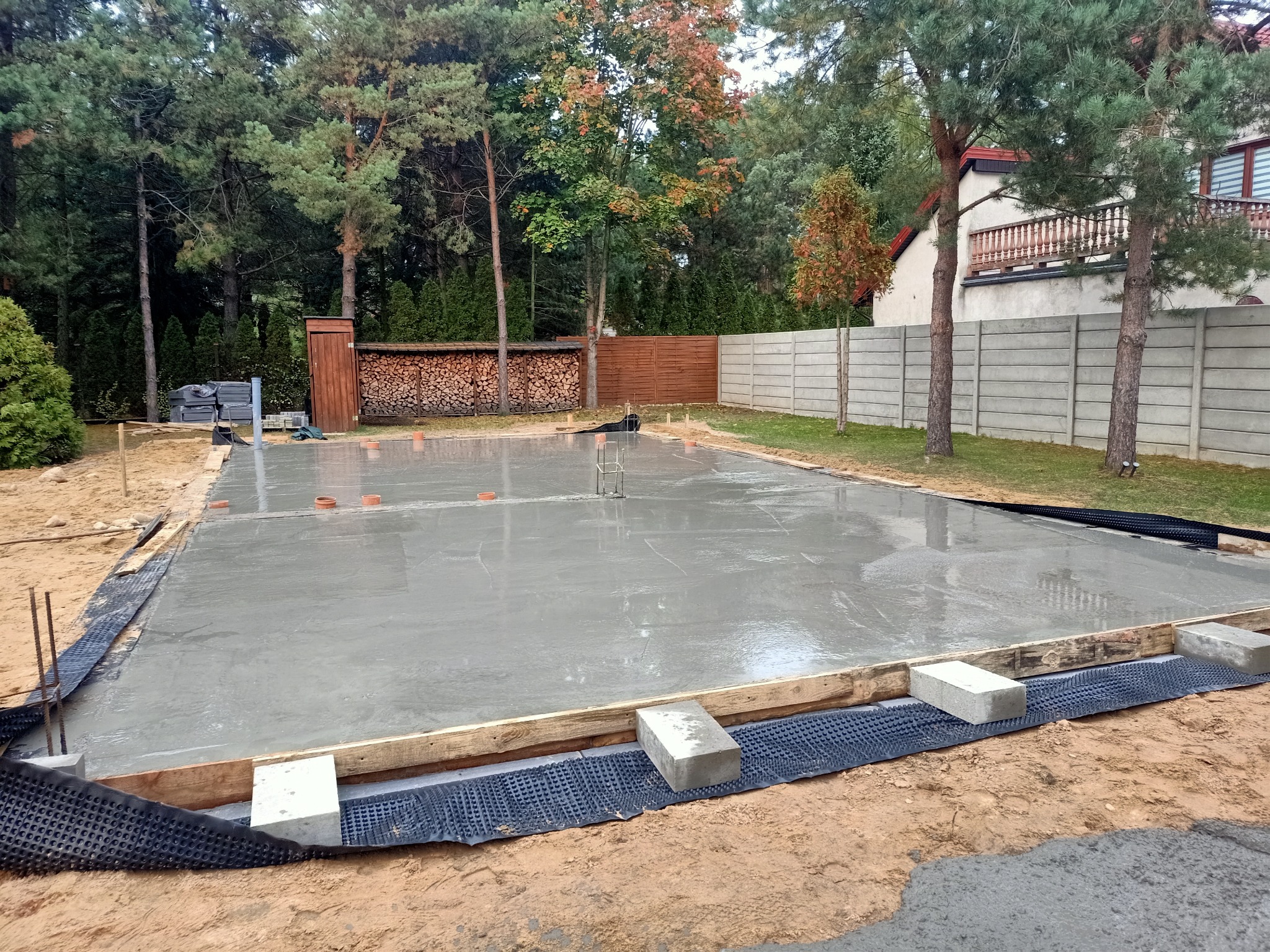 Świeżo wylany, mokry fundament pod budowę domu jednorodzinnego, widoczne szalunki, hydroizolacja pionowa i wystające pręty zbrojeniowe, w tle drewniana wiata na drewno i dom z balkonem.