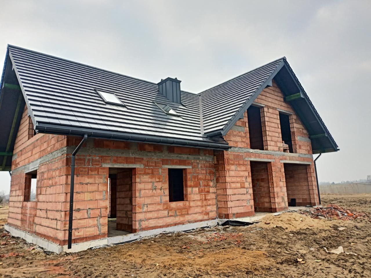 Surowy stan domu jednorodzinnego z czerwonej cegły z szarą dachówką, widoczne okna dachowe i orynnowanie, teren wokół w stanie budowy.