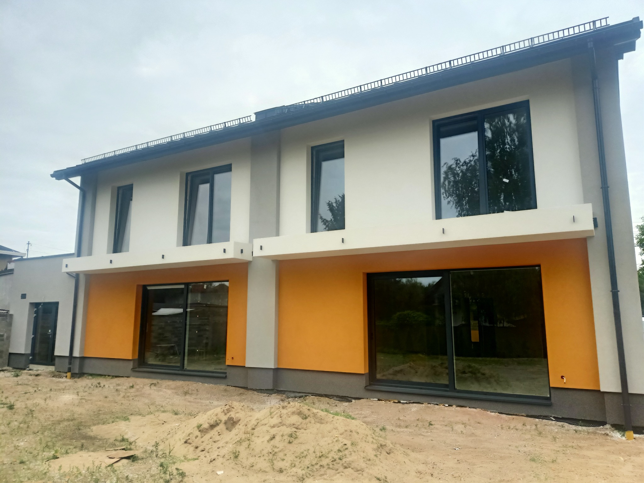 Nowoczesny, dwukondygnacyjny dom w zabudowie bliźniaczej z elewacją w odcieniach bieli, szarości i pomarańczy. Duże okna odbijają otoczenie. Widoczny fragment terenu przed budynkiem.