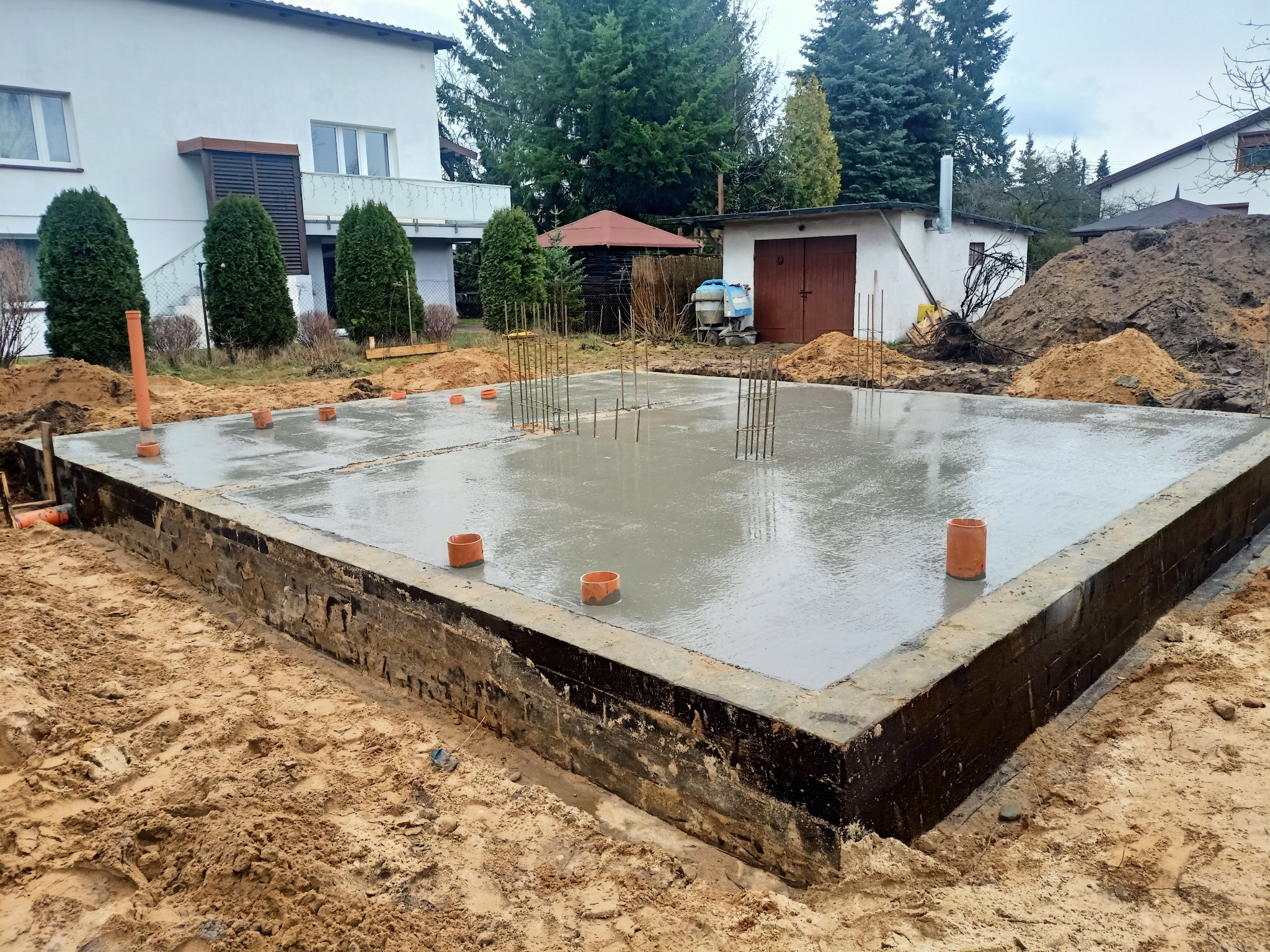 Świeżo wylany fundament domu jednorodzinnego z wystającymi prętami zbrojeniowymi, widoczne szalunki i rury wentylacyjne. W tle dom i sprzęt budowlany.