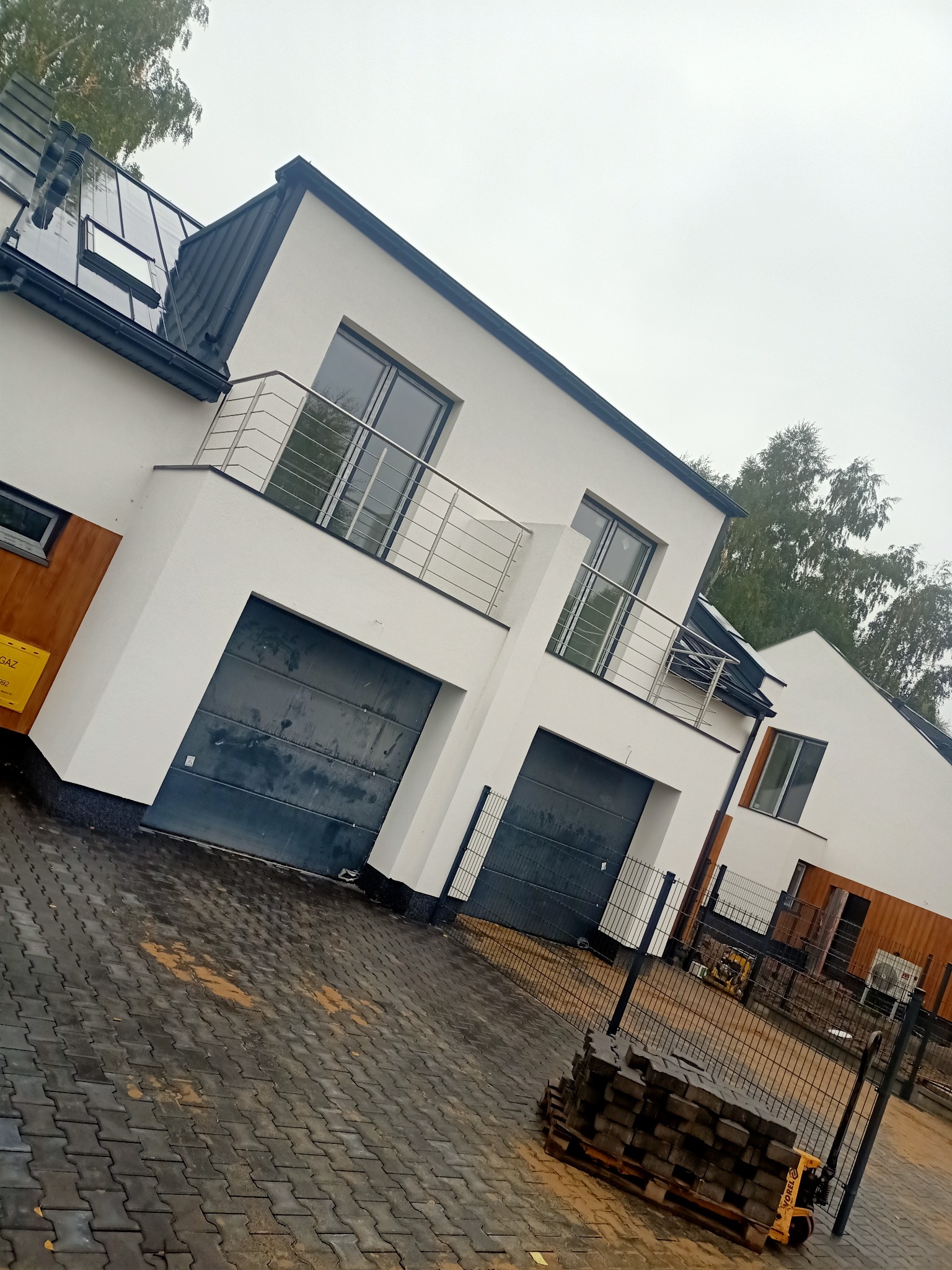 Nowoczesne domy szeregowe z balkonami i garażami, wykończone w bieli z elementami drewna, widok z zewnątrz na kostkę brukową i fragment placu budowy.