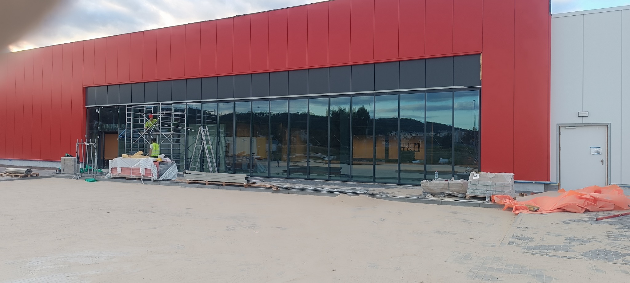 Montaż dużych, panoramicznych przeszkleń w budynku o czerwono-białej elewacji, widoczni pracownicy na rusztowaniu, palety z materiałami budowlanymi na placu budowy.