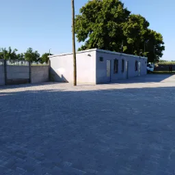 Rozległy plac wyłożony szarą kostką brukową, z budynkiem gospodarczym w tle i drzewem; słup energetyczny przechodzi przez dach budynku.