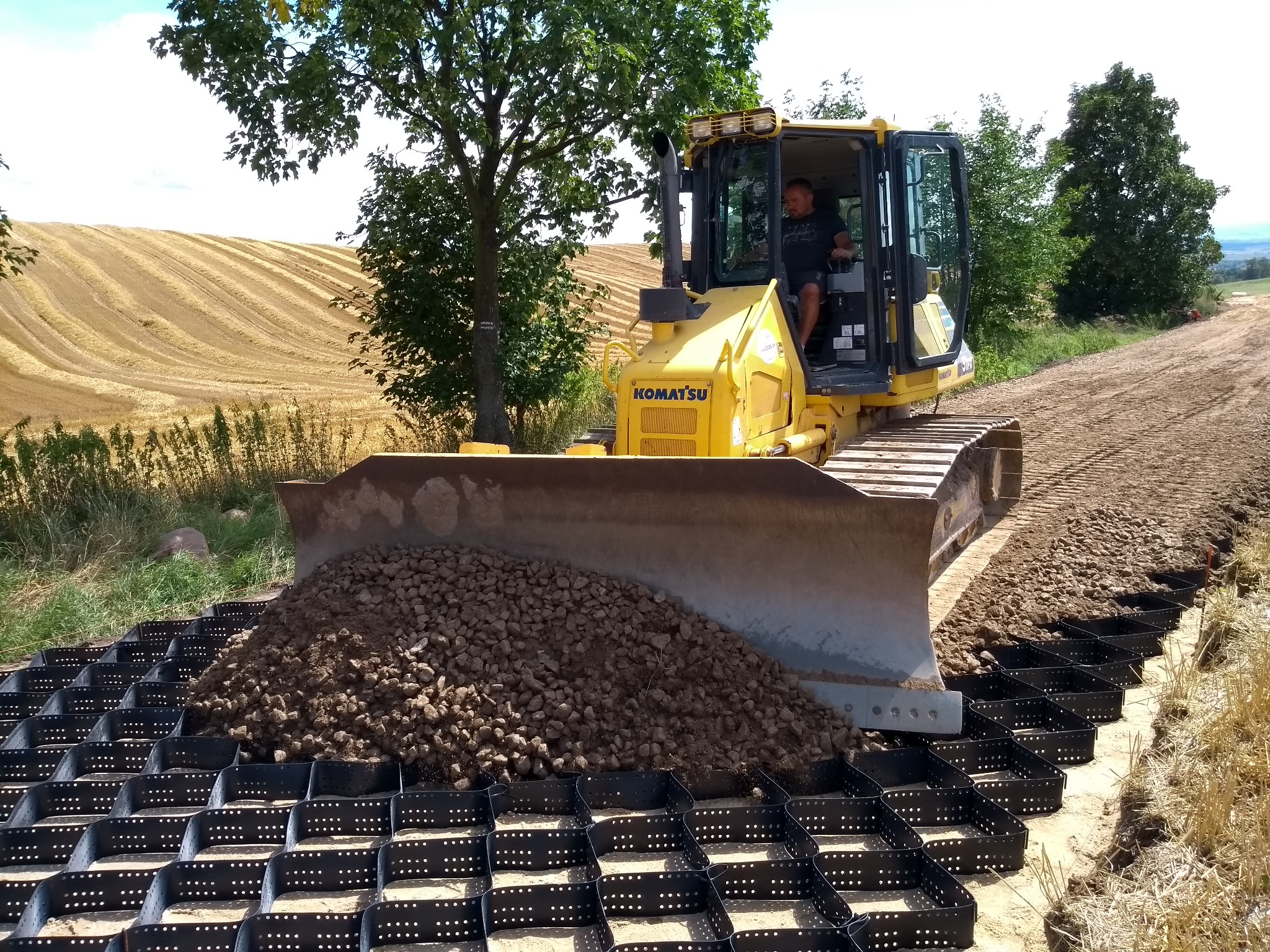 Żółty spychacz Komatsu z operatorem, nasypujący żwir na czarną geokratę drogową na tle pola i drzew, w słoneczny dzień.