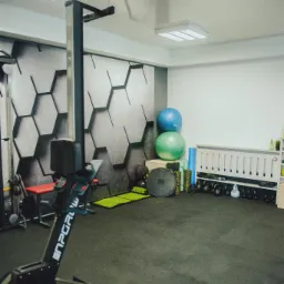 Wnętrze sali treningowej z czarną podłogą, ścianą z geometrycznym wzorem, sprzętem do ćwiczeń, takim jak wioślarz i kettlebells, oraz piłkami do ćwiczeń.