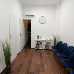 Logo.Pasja - Gabinet terapeutyczny JOANNA SIEREŃSKA - Jasne wnętrze poczekalni z niebieskimi krzesłami, drewnianą podłogą i białymi ścianami. Na ścianie zegar, a w tle widoczne drzwi do gabinetu.