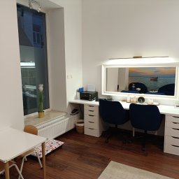 Logo.Pasja - Gabinet terapeutyczny JOANNA SIEREŃSKA - Jasne wnętrze gabinetu z biurkiem, lustrem z oświetleniem, dwoma krzesłami i stolikiem dla dzieci. W oknie widoczna zieleń, na biurku akcesoria.