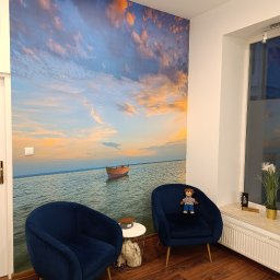 Logo.Pasja - Gabinet terapeutyczny JOANNA SIEREŃSKA - Wnętrze gabinetu z fototapetą przedstawiającą morze i łódkę, dwa granatowe fotele z pluszowym misiem na jednym z nich, drewniana podłoga.