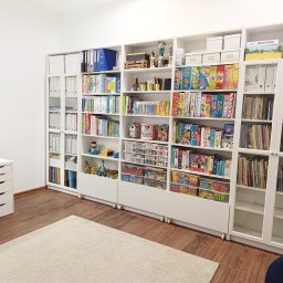 Logo.Pasja - Gabinet terapeutyczny JOANNA SIEREŃSKA - Jasny pokój z białymi regałami wypełnionymi książkami, grami i zabawkami. Biurko z krzesłem i dywan na podłodze. Uporządkowana przestrzeń terapeutyczna.