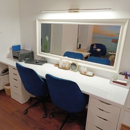 Logo.Pasja - Gabinet terapeutyczny JOANNA SIEREŃSKA - Jasne biuro z dwoma krzesłami i lustrem z oświetleniem. Na blacie biurka akcesoria biurowe i dekoracje. W lustrze odbija się pokój z kanapą i obrazem.